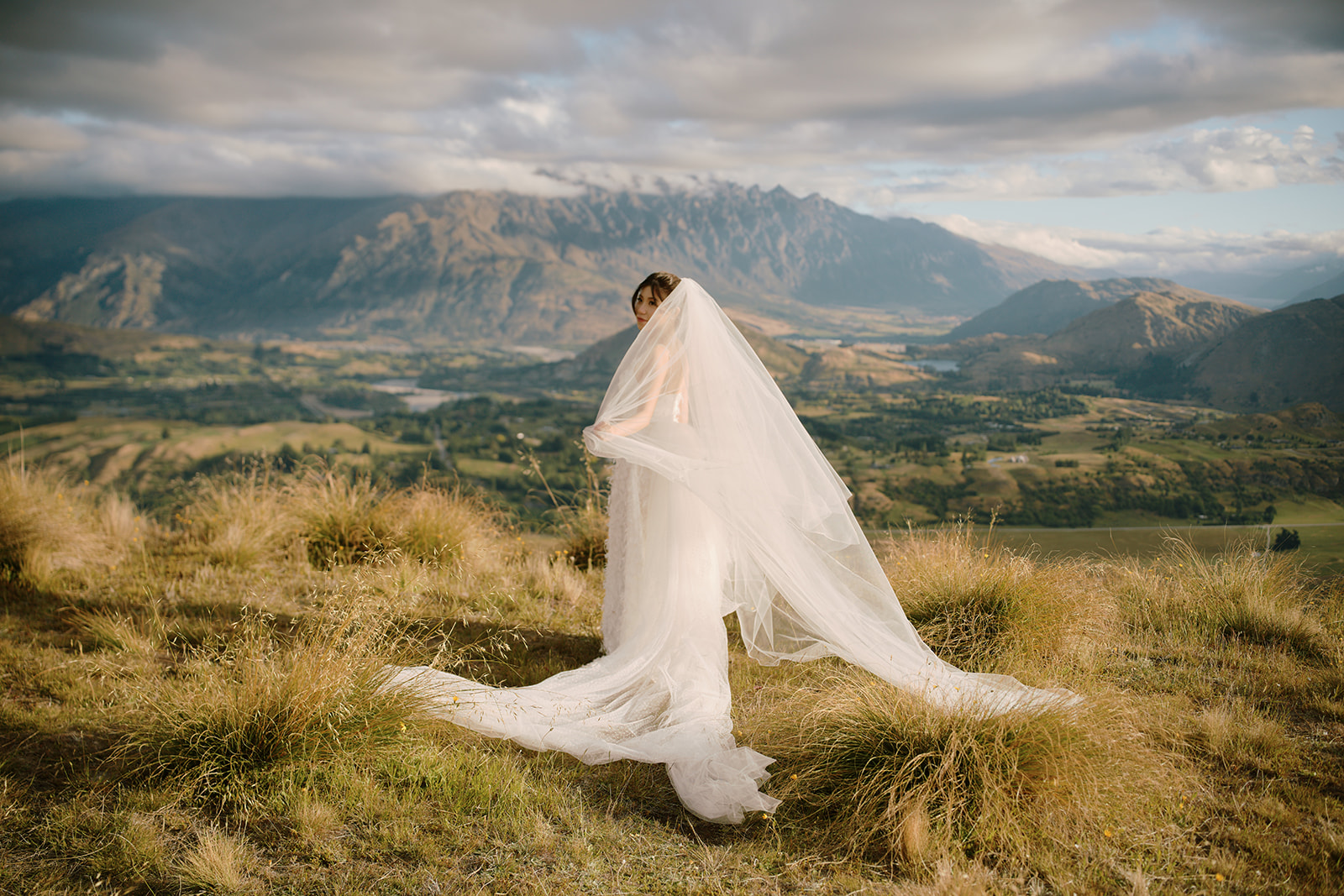 クイーンズタウン　ニュージーランド　ウェディング　エロープメント　前撮り　フォト　結婚写真 Queenstown Wedding Photographer A bride is standing on top of a hill in ニュージーランド with mountains in the background.